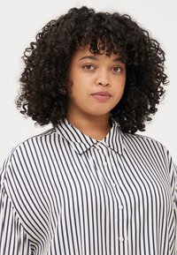 Ung kvinne med krøllete hår iført en stripete skjorte med knapper. Hun poserer med et nøytralt uttrykk mot en ensfarget bakgrunn.