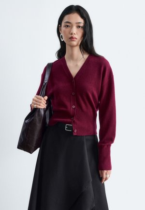 Jeune femme aux longs cheveux noirs porte un cardigan bordeaux boutonné, une jupe noire, et porte un sac en cuir sombre à l'épaule.