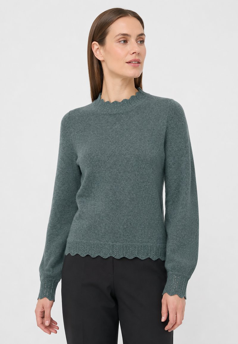 Femme aux cheveux bruns et lisses portant un pull en tricot vert à bords festonnés et un pantalon noir, regardant sur le côté sur un fond uni.
