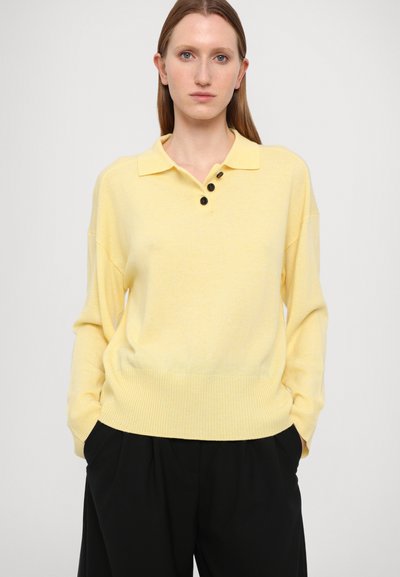Suéter amarillo hecho de una tela suave con un cuello y tres botones negros. Presenta un dobladillo acanalado y un corte relajado, combinado con pantalones negros.