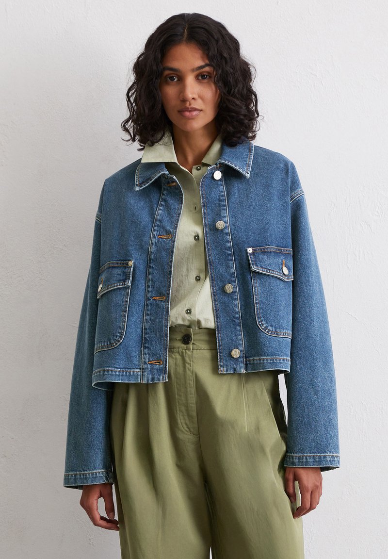 Kobieta z kręconymi włosami ma na sobie krótką dżinsową kurtkę na jasną zieloną koszulę i oliwkowe spodnie, stojąc na neutralnym tle.