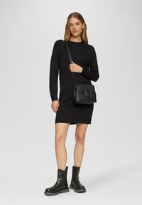 Schwarzes Strickkleid mit langen Ärmeln, gerippten Bündchen und lockerer Passform. Kombiniert mit schwarzen Stiefeletten und einer strukturierten Umhängetasche.