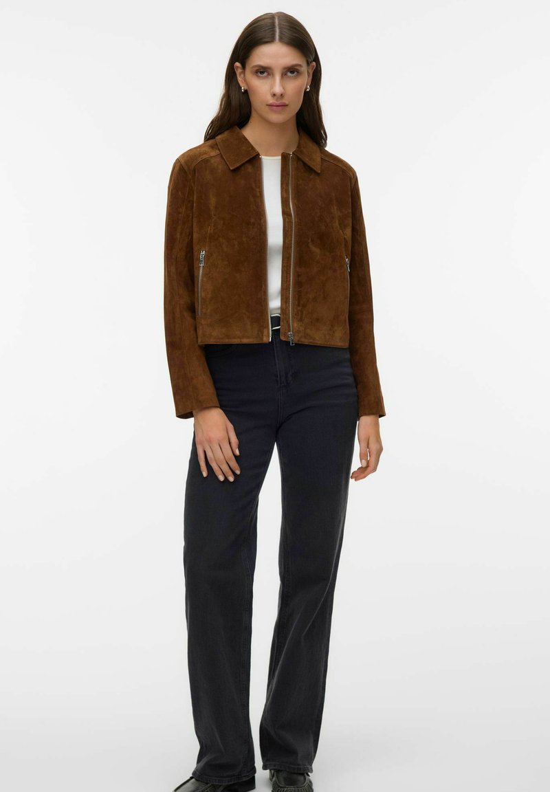 Femme aux longs cheveux bruns portant une veste zippée en daim marron, un haut blanc, un jean noir et des chaussures noires, posant devant un fond blanc.