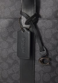Black leather bag with textured fabric, featuring a hang tag, decorative strap, and C-shaped hardware accent. Subtle pattern detail visible.