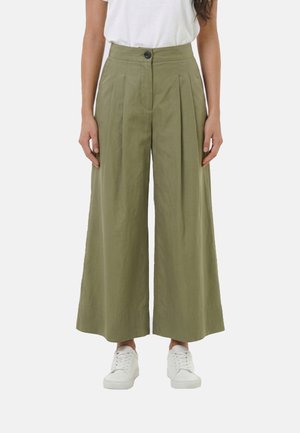 Donna con pantaloni a vita alta, larghi e olive con pieghe e bottone frontale, indossa sneakers bianche, in piedi davanti a uno sfondo semplice.