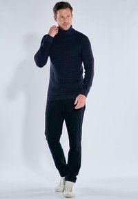 Mann, der den Kragen eines marineblauen Rollkragenpullovers anpasst, trägt dunkle Hosen und weiße Sneaker, steht vor einem schlichten hellen Hintergrund.