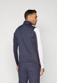 Marineblå zip-up vest med høy krage, hvite lange ermer og burgunder side-striper. Glatt stoff med atletisk passform og minimalistisk design.
