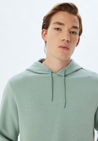 Sudadera con capucha verde menta de textura suave, con un bolsillo tipo canguro y detalles de cordón. Diseño simple, tejido sin adornos.