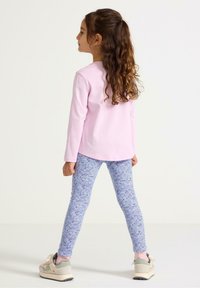 Långärmad rosa skjorta, blommönstrade blå leggings och beige sneakers med grå detaljer. Barnet står med ryggen vänd mot betraktaren.