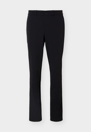 Modelo masculino de pie con una camiseta polo beige de manga corta, pantalones negros con cinturón y zapatillas blancas sobre un fondo claro y liso.