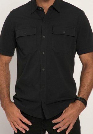 Mann trägt ein schwarzes kurzärmeliges Hemd mit zwei aufgesetzten Taschen vorne und ein schwarzes Armband am linken Arm.