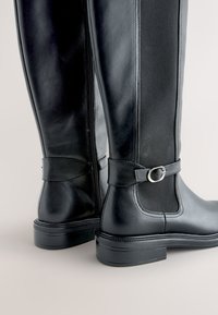 Schwarze kniehohe Lederstiefel mit seitlichem Reißverschluss, elastischem Einsatz, Riemen und silberner Schnalle, mit niedrigem Blockabsatz und abgerundetem Zehenbereich.