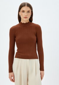 Maglione a collo alto marrone a coste con maniche lunghe e scollatura ondulata, abbinato a pantaloni plissettati color crema.