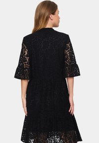 Black lace dress featuring a round neckline, bell sleeves, and a tiered hem. Delicate floral pattern throughout adds texture.