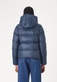 Personne portant une doudoune bleu marine avec une capuche et un jean bleu, se tenant de dos devant un fond uni.