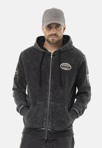 Hoodie gris zippé en tissu texturé, avec une capuche, des poches avant et des patches logo brodés sur les manches et la poitrine.
