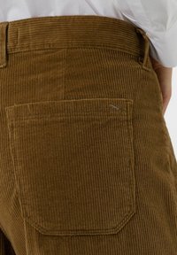Brązowe spodnie sztruksowe z pionowymi prążkami, kieszenią naszywaną i subtelnymi detalami w szwie. Gładka tekstura, dopasowany krój.