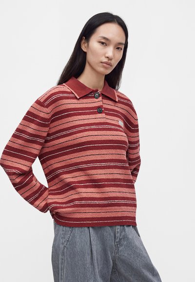 Femme aux longs cheveux foncés portant un pull boutonné rayé rouge et rose ainsi qu'un pantalon gris ample sur un fond clair uni.