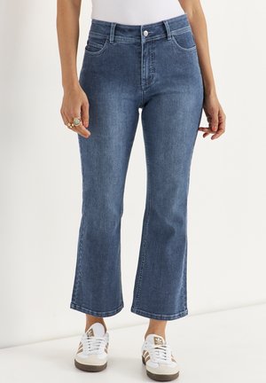 Vrouw draagt blauw, licht uitlopende jeans met een mid-rise taille, voorzakken en witte sneakers met bruine strepen.