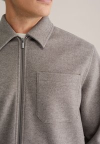 Lichtgrijze wollen zip-up jas met een puntige kraag, voorzak en metalen rits. Gladde textuur, minimalistisch ontwerp.