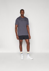 Graues Kurzarm-T-Shirt mit schwarzem Logo, kombiniert mit schwarzen Shorts und weißen Turnschuhen. Enthält gestreifte Socken mit blauen und gelben Akzenten.