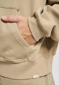 Hoodie beige en tissu doux, avec une poche frontale, des poignets côtelés et une petite étiquette de marque sur l'ourlet.
