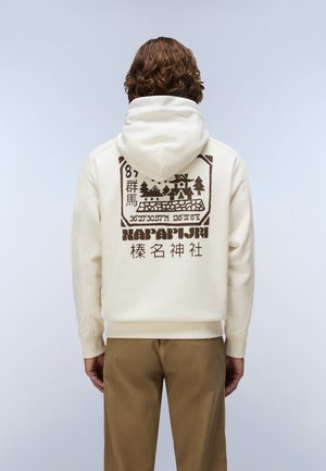 Persoon met krullend haar die een crèmekleurige hoodie draagt met een bruine afbeelding van een huis, bomen, coördinaten en Japanse tekst op de achterkant.