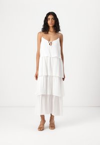 Robe maxi blanche à volants avec des fines bretelles et un détail noué à l'avant. Fabriquée en tissu léger et texturé. Associée à des sandales à lanières.