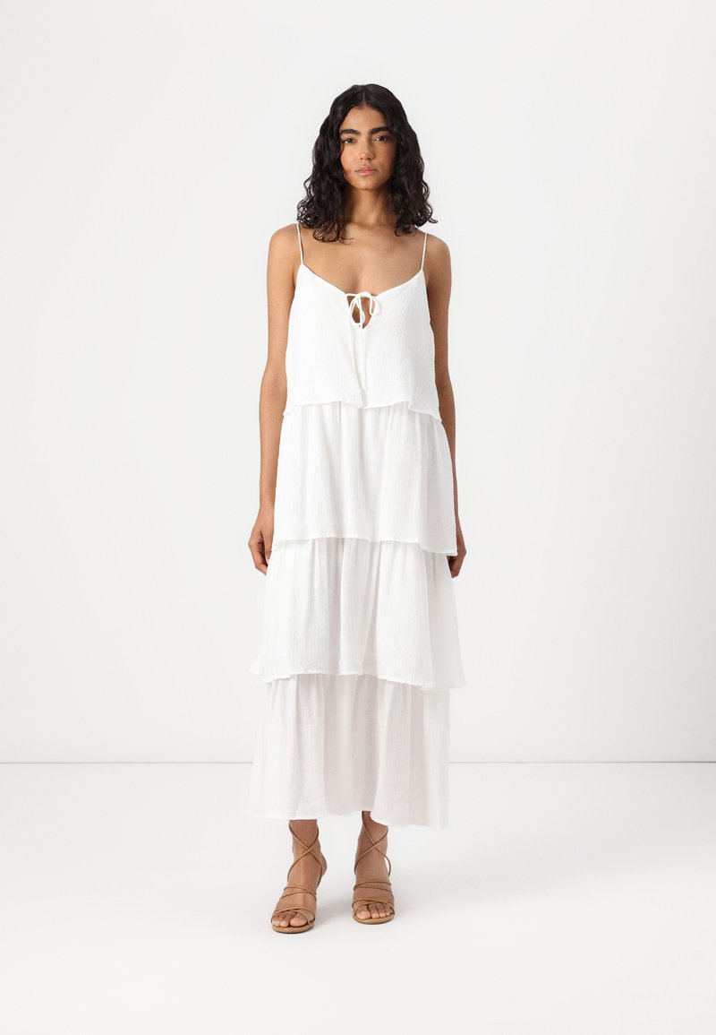 Robe maxi blanche à volants avec des fines bretelles et un détail noué à l'avant. Fabriquée en tissu léger et texturé. Associée à des sandales à lanières.