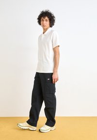 Polo shirt blanc à manches courtes, pantalon cargo bleu marine avec poches latérales, et baskets vert clair avec une semelle texturée. Design et couleurs simples.