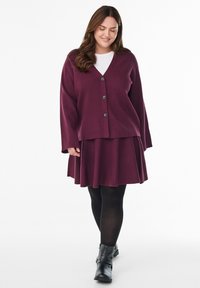 Žena v švestkovém cardigan s knoflíky a odpovídající sukni, s černými punčocháči a černými kotníkovými botami, stojící na bílém pozadí, usmívající se.