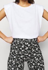 Vit bomullströja med korta, strukturerade ärmar som bärs med svarta byxor med blommönster. Tygmaterialet har en texturerad, lätt finish.