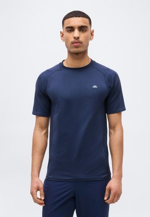 Mladý muž stojaci, oblečený v tmavomodrom športovom tričku s krátkym rukávom a zhodných tmavomodrých nohaviciach na jednoduchom svetlom pozadí.