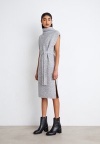 Vestido gris de punto con cuello alto, mangas cortas y cintura ajustada con lazo. Aberturas laterales y combinado con botas negras hasta el tobillo con tacones cuadrados.