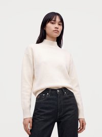 Junge Person mit langen dunklen Haaren, die einen cremefarbenen Rollkragenpullover und hoch taillierte schwarze Jeans trägt, vor einem schlichten hellen Hintergrund.