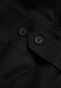 Schwarzer Stoff mit strukturierter Oberfläche hat zwei schwarze Knöpfe. Das Design beinhaltet eine Klappe mit Stichdetail und eine Taschenöffnung.