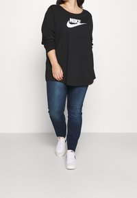 Svart långärmad Nike-tröja med vitt logo, kombinerat med mörkblå jeans och vita sneakers, stående mot en neutral bakgrund.