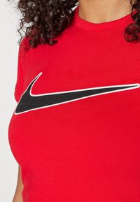 Czerwona bawełniana koszulka z dużym czarnym logo Nike Swoosh obramowanym białym kolorem, dopasowany krój i krótkie rękawy.