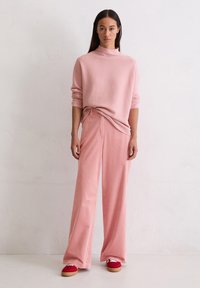 Roze gebreide trui met een hoge hals, gecombineerd met wijde roze corduroy broek. Aangevuld met rode schoenen en minimalistische designdetails.