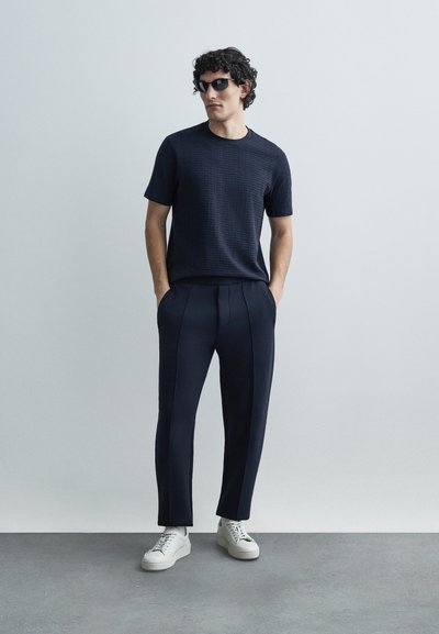 Tmavomodrá textúrovaná košeľa s krátkym rukávom, tmavomodré nohavice so zúženými nohavicami a prednými švami, a biele tenisky. Oblečenie dopĺňajú slnečné okuliare.