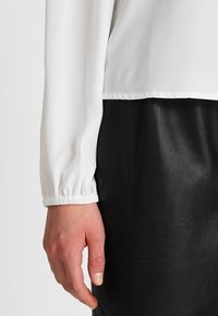 Vit långärmad topp med ärmslut som är rynkade, kombinerad med en svart skinnkjol. Mjuk tyg, slät textur och en avslappnad passform är tydligt synliga.