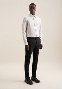 Chemise blanche à boutons avec de petits pois, assortie à un pantalon noir et des chaussures noires. Le mannequin se tient devant un fond neutre.