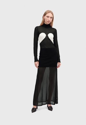 Femme aux longs cheveux blonds portant une longue robe noire transparente avec des formes de cœurs blancs sur la poitrine, chaussures noires pointues, debout sur un fond clair.