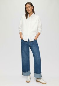 Witte blouse met lange mouwen, gecombineerd met een wijde blue jeans en witte sneakers met beige accenten en opgerolde manchetten.