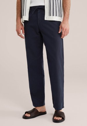 Bărbat purtând pantaloni bleumarin cu șnur, cămașă albă, cardigan cu model, inel argintiu și sandale împletite maro închis.