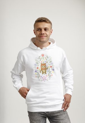 Junger Mann trägt weißen Hoodie mit Cartoon-Ewok, der einen Speer hält, umgeben von bunten Blumenmustern, steht vor einfachem Hintergrund.