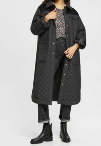 Cappotto lungo imbottito nero con collo a camicia, bottoni a pressione, tasche laterali e una superficie liscia, abbinato a jeans scuri e stivali alla caviglia.