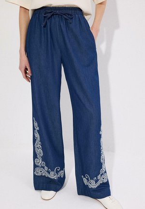 Pantalones anchos azules con patrones de cachemira bordados en blanco cerca del dobladillo, cintura elástica y cordón, usados con zapatos blancos.