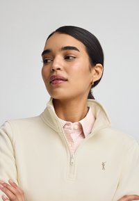 Fleece crema con zip e colletto alto, indossato sopra una camicia a scacchi rosa chiaro. Presenta un piccolo logo ricamato sul petto.