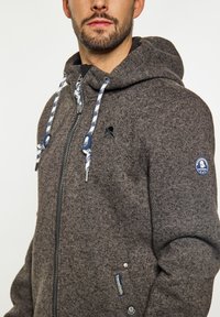 Sivi zip-up dukserica s teksturiranom tkaninom, kontrastnim plavim i bijelim uvećanjima, te logom na prsima i rukavu.
